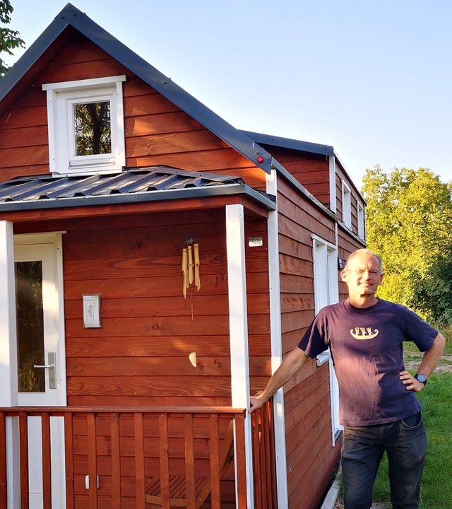 "Terra Xpress: Mietest Du noch oder baust Du schon?": Oliver Victor steht vor einem seiner braunen Tiny-Houses und lehnt sich an den Zaun der kleinen Veranda.