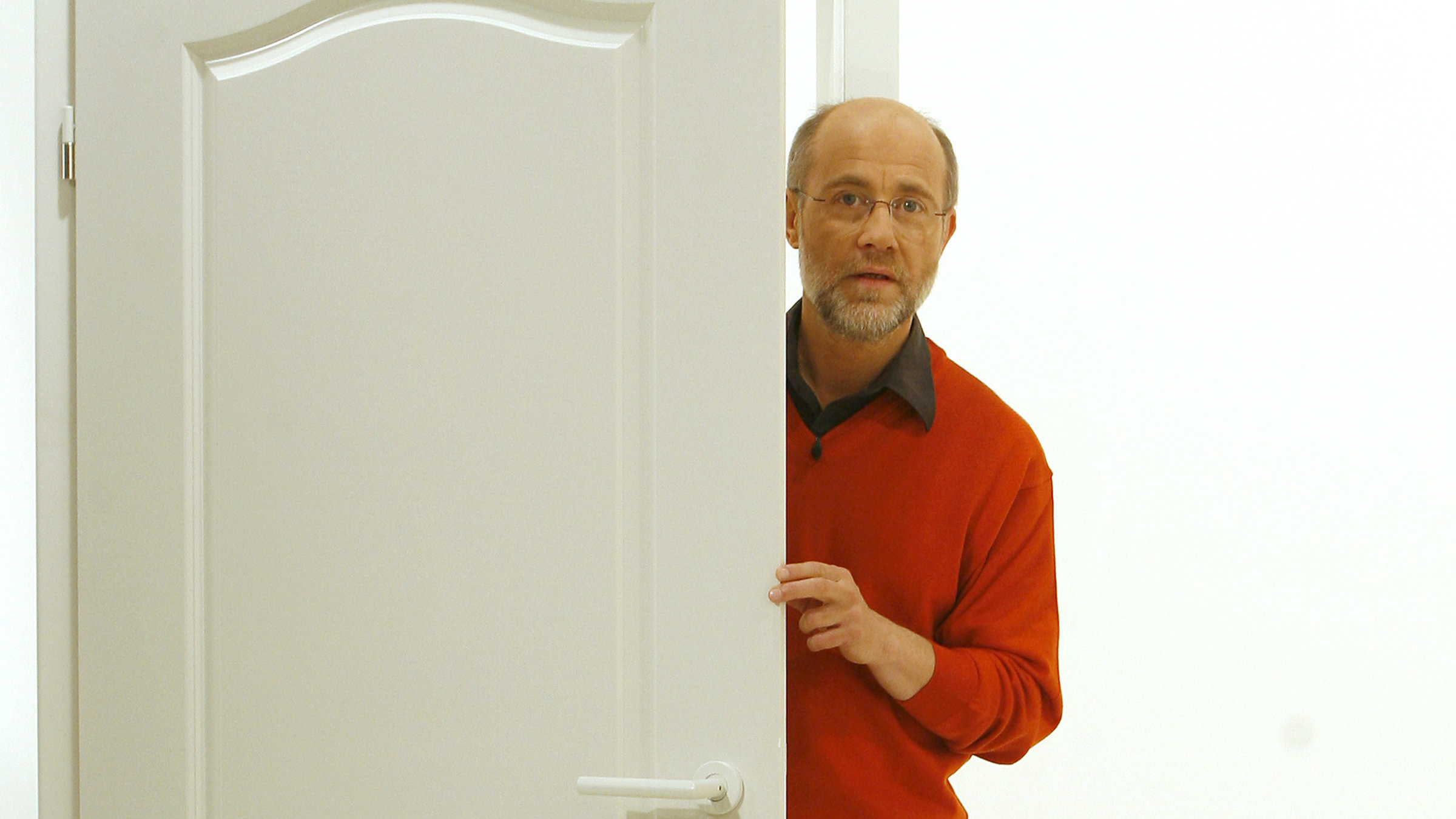 "Frag den Lesch": Prof. Harald Lesch steht - in rotem Pulli und schwarzem Hemd - in einer weißen, halb geöffneten Tür und blickt freundlich in Richtung Kamera.
