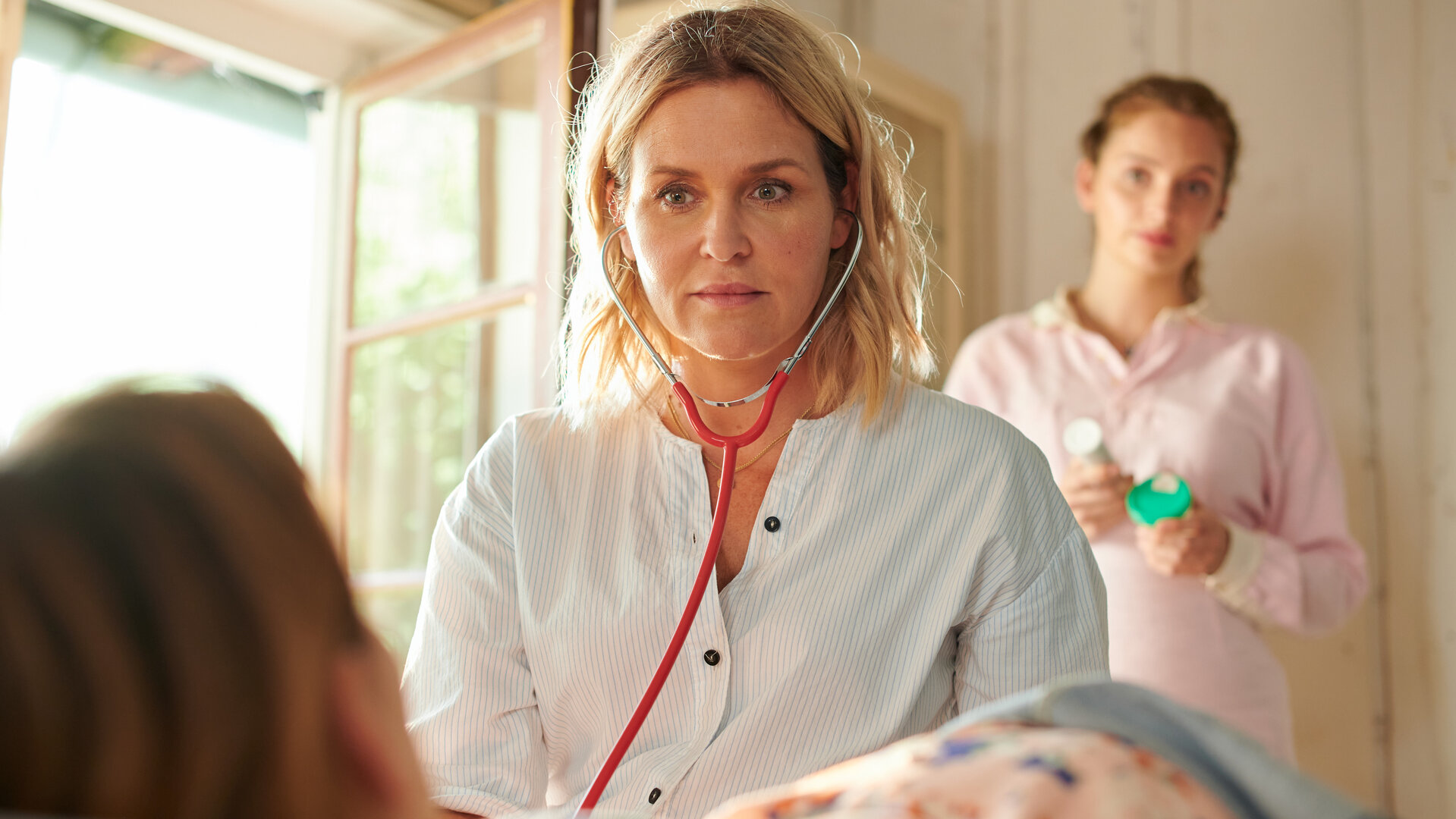"Lena Lorenz - Krank vor Sorge": Lena Lorenz (Judith Hoersch) untersucht die schwangere Tati Meisinger (Anouk Elias), die auf einer Liege Platz genommen hat und nur von hinten zu sehen ist. Im Hintergrund beobachtet die angehende Hebamme Celine Gollert (Paulina Rümmelein) das Geschehen aufmerksam.