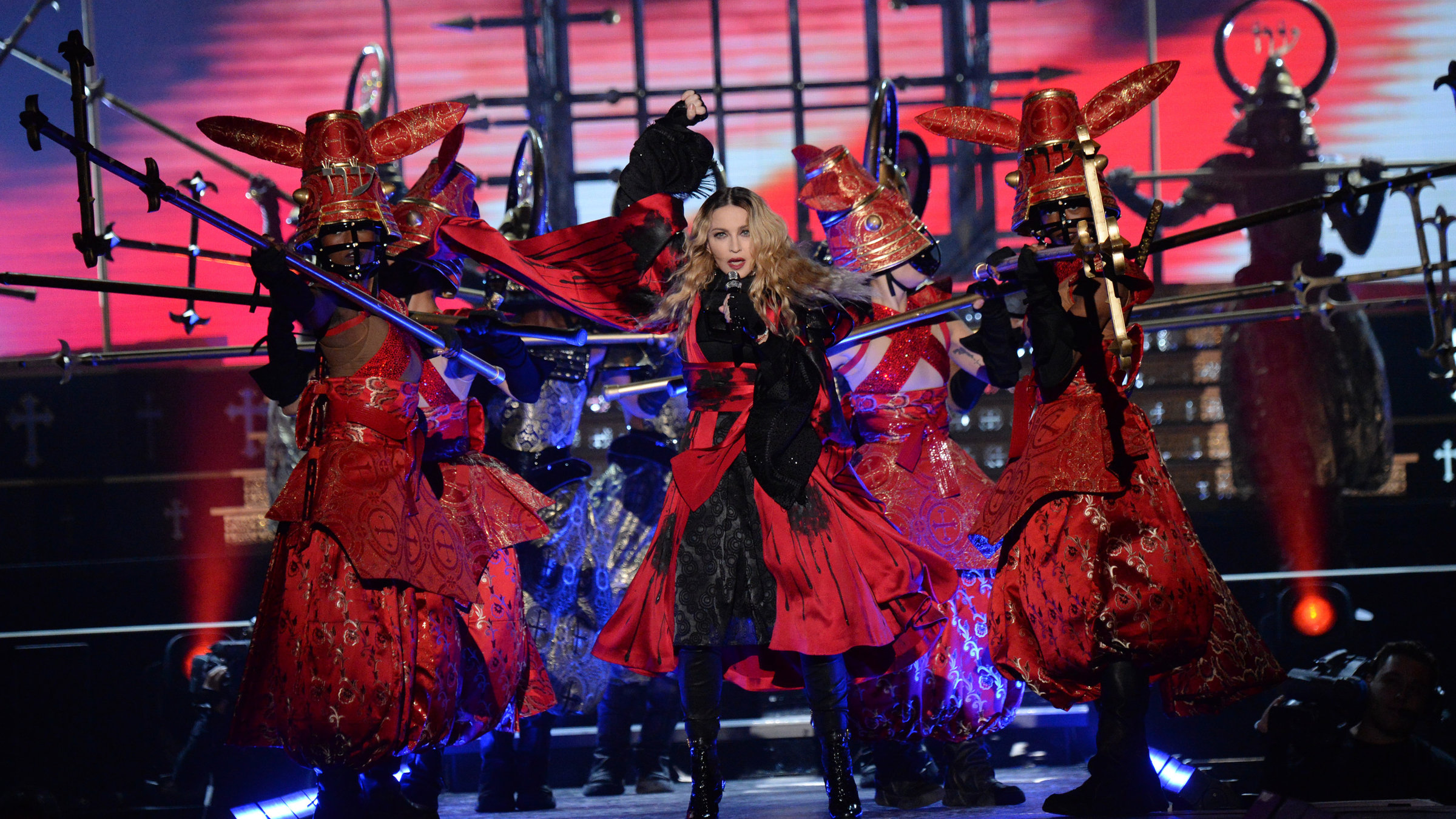 "Madonna: Rebel Heart Tour:" Die Sängerin Madonna auf der Bühne. Sie trägt eine rot-schwarzes Kleid, schwarze fingerlose Handschuhe und schwarze Lackstiefel. In der linken Hand hält sie ein Mikrofon. Hinter ihr mehrere Tänzerinnen in roten Kleidern und mit roten auffälligen Kopfbedeckungen.