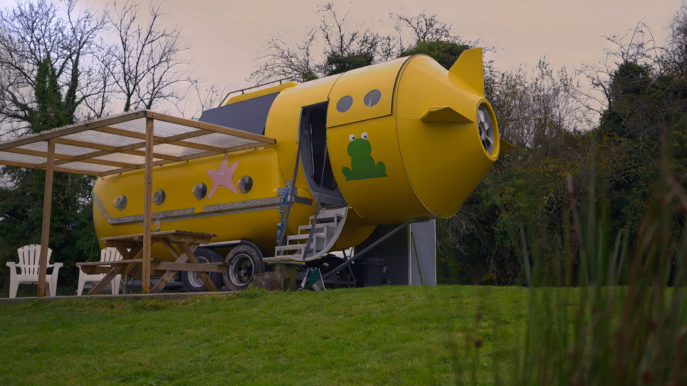 "Die außergewöhnlichsten Hotels der Welt - Versteckte Rückzugsorte": Gelbes Ufo-förmiges Wohnmobil mit Frosch-Aufkleber und Stern, auf einem Anhänger stehend, umgeben von Bäumen und Wiese.