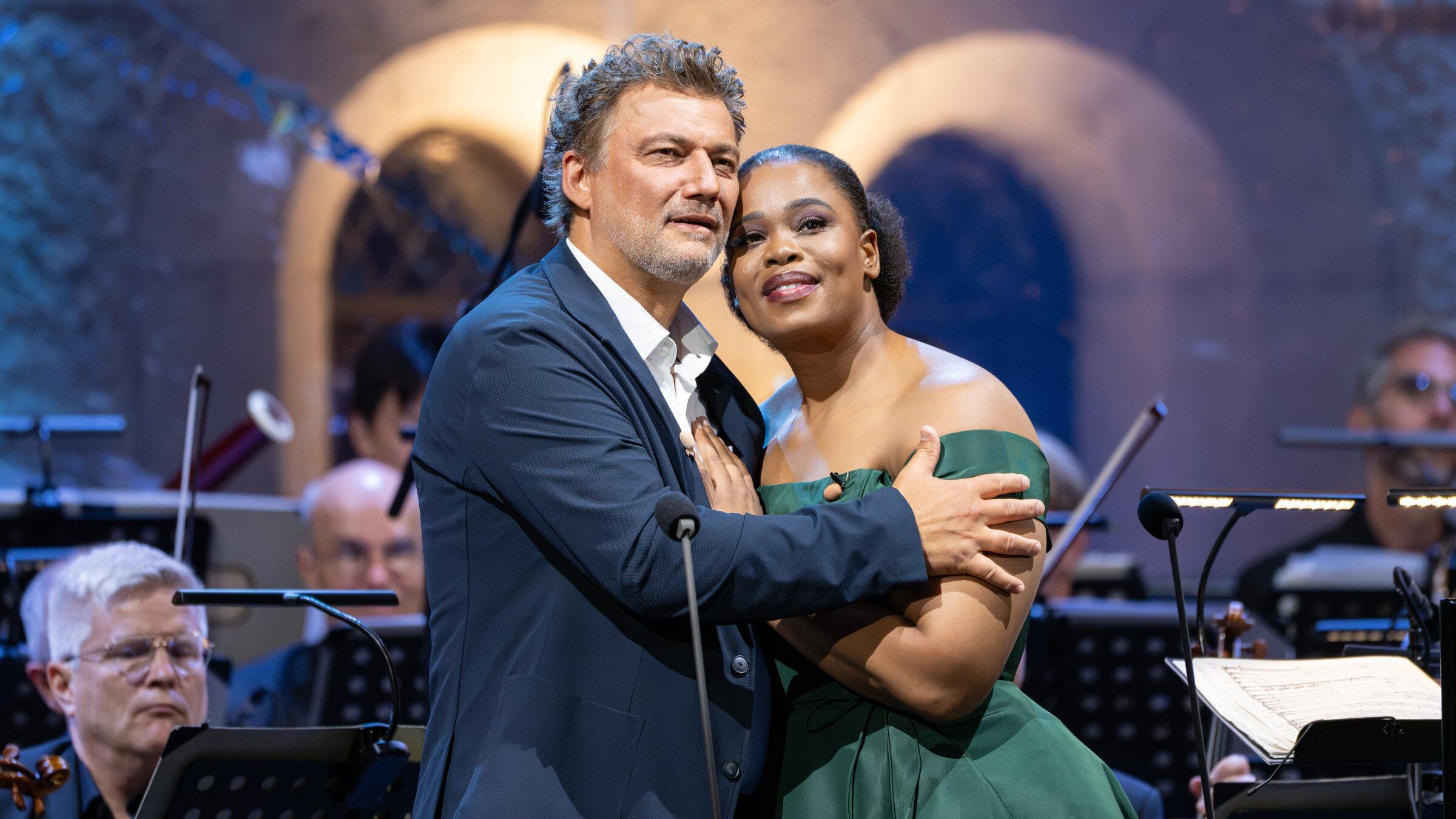 "Oper unter Sternen - mit Jonas Kaufmann und Pretty Yende auf Schloss Neuschwanstein": Jonas Kaufmann und Pretty Yende stehen auf einer BÃ¼hne. Sie umarmen sich und blicken vertrÃ¤umt in die Kamera. Im Hintergrund sitzen die Orchestermusiker.