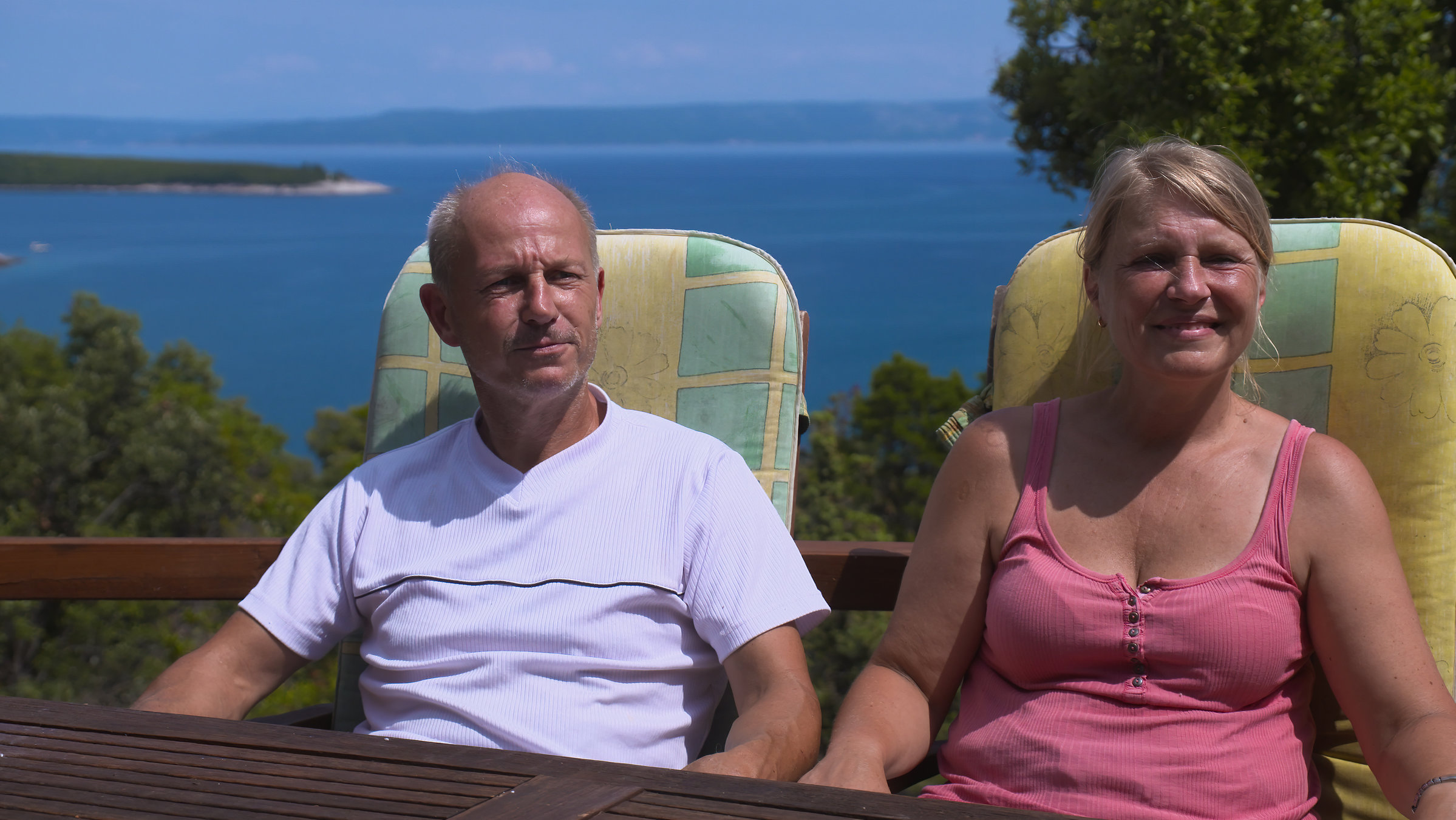 "37°: Späte Träume, neue Welten - Auswandern im Ruhestand": Ralf (53) und Sylvia (51) sitzen auf Gartenstühlen und schauen an der Kamera vorbei. Hinter ihnen liegt das Meer.