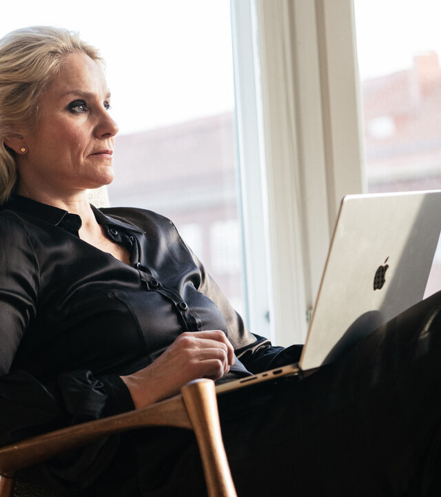 "Queen of Fucking Everything - Zahltag": Linda (Laura Malmivaara) sitzt in einem Raum vor einer Fensterfront. Auf ihrem Schoß steht ein aufgeklappter Laptop.