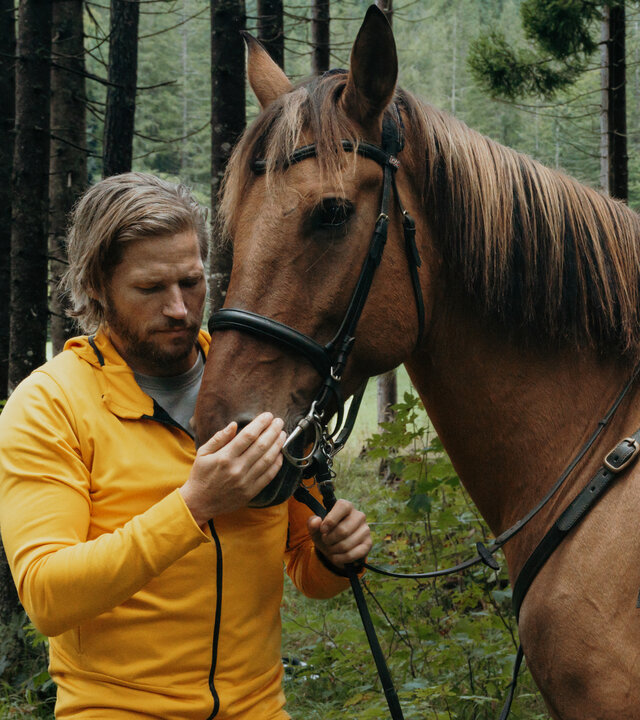 "Die Bergretter: Gespenster": Im Wald: Bergretter Markus Kofler (Sebastian Ströbel) steht am Kopf eines braunen Pferds, das gesattelt ist. Mit einer Hand hält er das Zaumzeug fest, die andere Hand hat er auf das Maul gelegt. Er wirkt beruhigend auf das Tier ein.