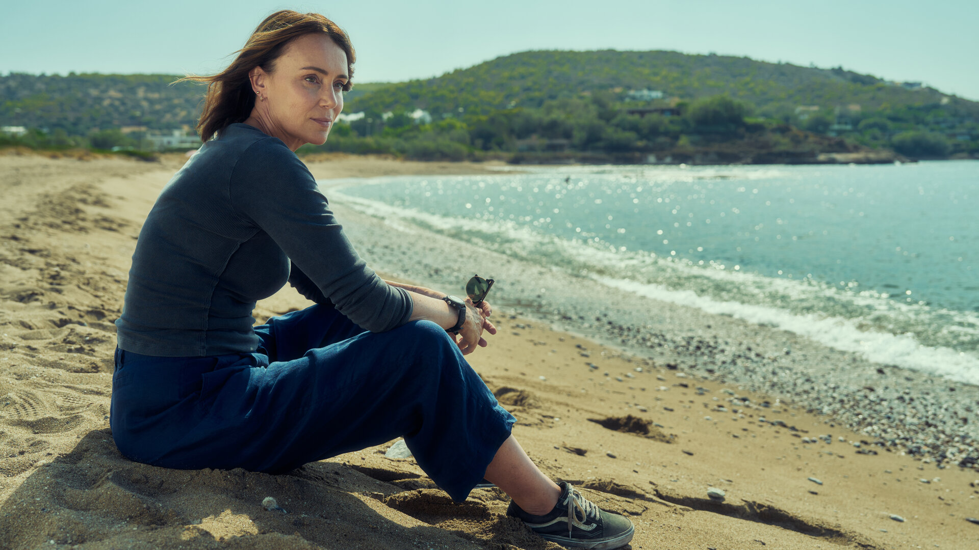 "The Assassin - Eine explosive Hochzeit": Julie Green (Keeley Hawes) sitzt an einem Strand am Meer. Im Hintergrund liegt eine hügelige Landschaft mit vielen Bäumen.