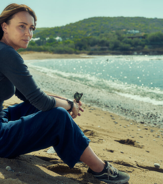 "The Assassin - Eine explosive Hochzeit": Julie Green (Keeley Hawes) sitzt an einem Strand am Meer. Im Hintergrund liegt eine hügelige Landschaft mit vielen Bäumen.