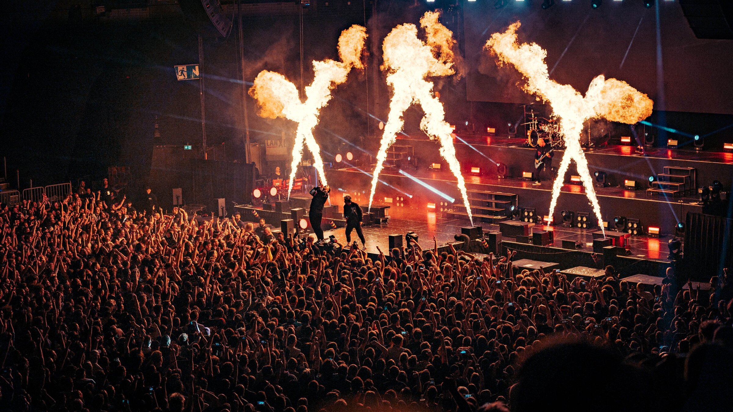 "Electric Callboy: Tekkno Tour '23 - Live in Europe": Blick auf die Band auf einer aufwendig gestalteten Bühne mit unterschiedlichen Ebenen, Licht- und Pyrotechnik-Effekten. Davor steht dichtgedrängt das Publikum und streckt teils die Arme in die Luft.