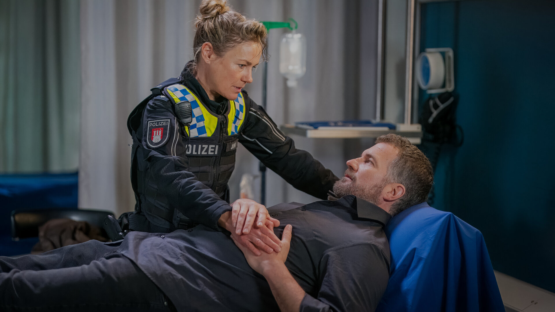 "Notruf Hafenkante - Schuld und Sühne": Philipp (Simon Böer) liegt in einem Behandlungsraum des EKH auf einer Patientenliege. Neben ihm steht die Polizistin Franzi Jung (Rhea Harder-Vennewald). Sie hat ihre rechte Hand auf Philipps Bauch zwischen seinen Händen liegen. Beide sehen sich an.
