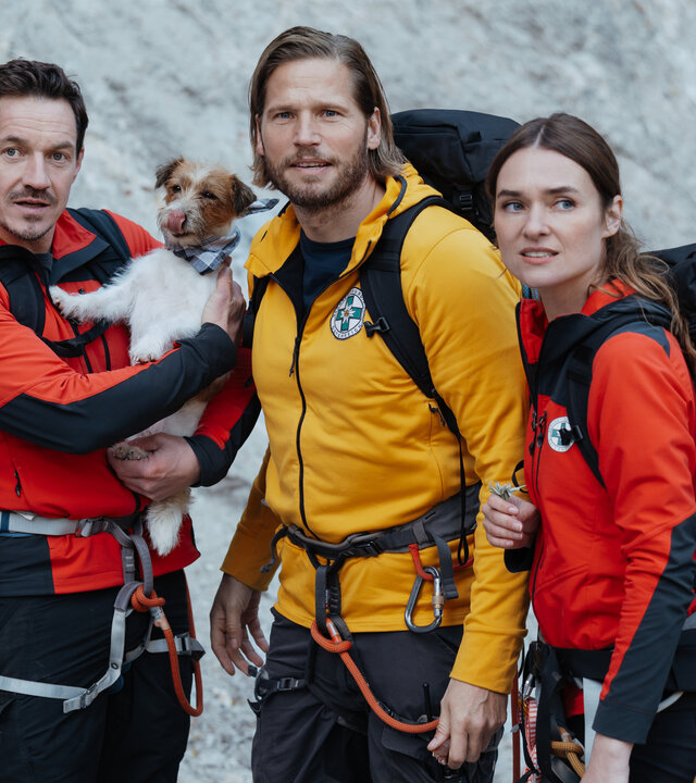 "Die Bergretter: Spiel mit dem Feuer": Vor einer Felswand stehen die Bergretter Tobi (Markus Brandl), Markus (Sebastian StrÃ¶bel) und Nina (Josephin Busch)nebeneinander und blicken auf etwas auÃerhalb des Bildrands. Sie tragen neben ihren RÃ¼cksÃ¤cken ihre RettungsausrÃ¼stung. Tobi hÃ¤lt einen kleinen Hund auf dem Arm. Dieser trÃ¤gt ein Halsband und leckt sich mit der Zunge Ã¼ber die Nase.