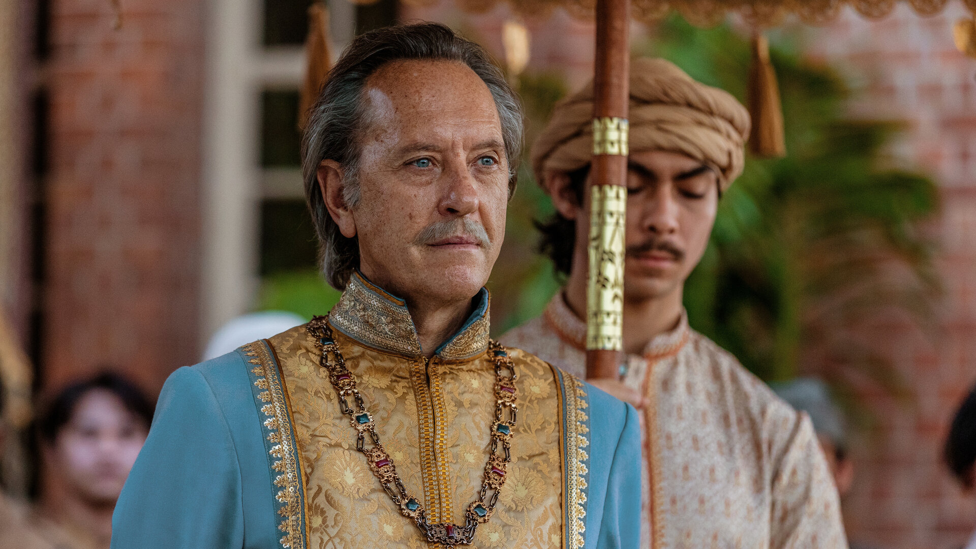 "Nautilus - Der Krake": Der Radscha (Richard E. Grant) steht vor einem Gebäude. Er ist farbenfroh gekleidet und trägt eine große, mit Edelsteinen verzierte Kette um den Hals. Hinter ihm hält ein Diener (Komparse) einen Sonnenschirm, von dem nur der Stock zu sehen ist. Im Hintergrund sind unscharf weitere Menschen erkennbar.