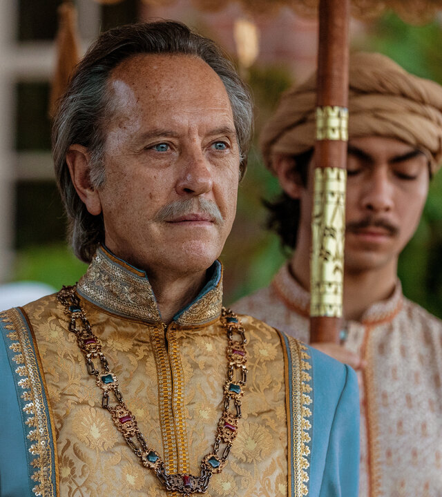 "Nautilus - Der Krake": Der Radscha (Richard E. Grant) steht vor einem Gebäude. Er ist farbenfroh gekleidet und trägt eine große, mit Edelsteinen verzierte Kette um den Hals. Hinter ihm hält ein Diener (Komparse) einen Sonnenschirm, von dem nur der Stock zu sehen ist. Im Hintergrund sind unscharf weitere Menschen erkennbar.