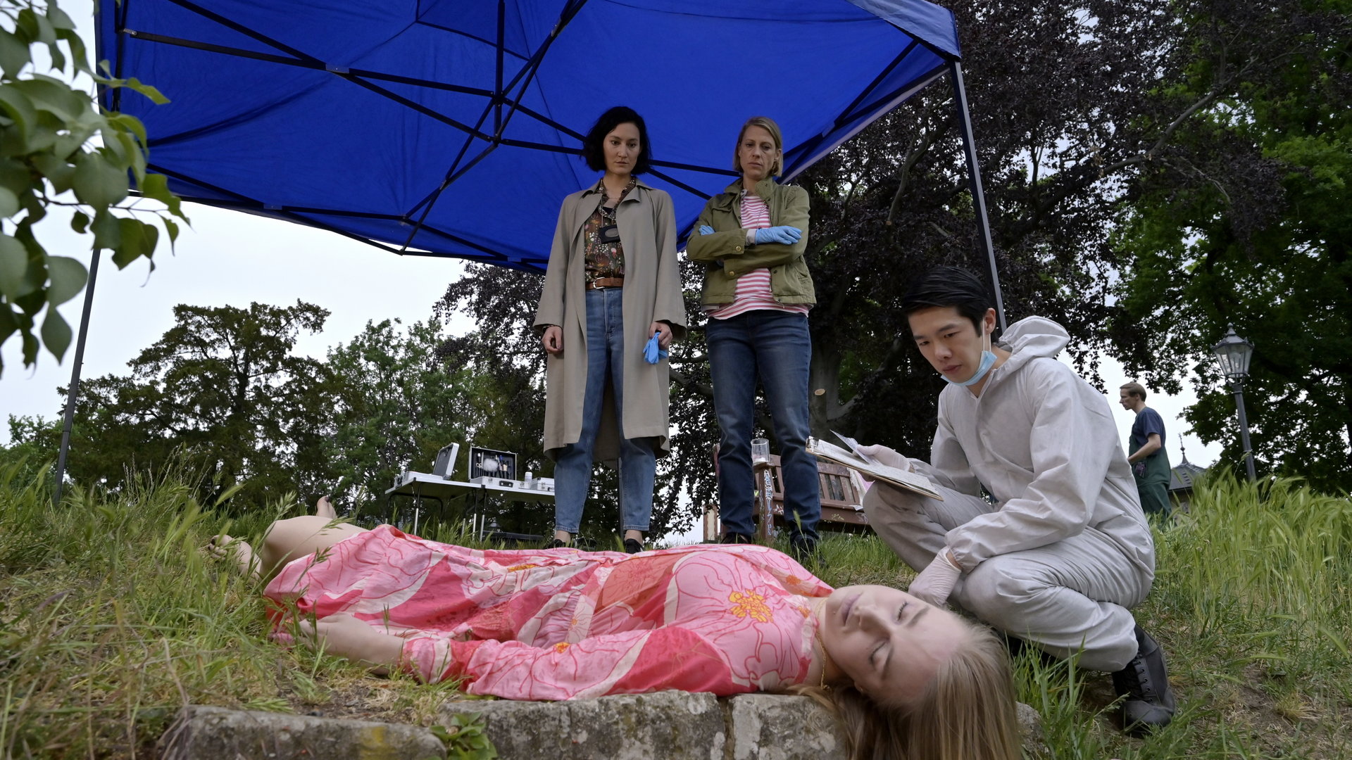 "SOKO Potsdam - Das siebte Haus": Auf einer Wiese auf dem Pfingstberg liegt eine weibliche Leiche, vor der Spurensicherer Thomas Brandner (Yung Ngo) hockt und die Leiche inspiziert. Pauline Hobrecht (Agnes Decker) und Tamara Meurer (Anja Pahl) stehen vor der Leiche unter einem blauen Sonnenschirm und blicken hinunter auf die Leiche.