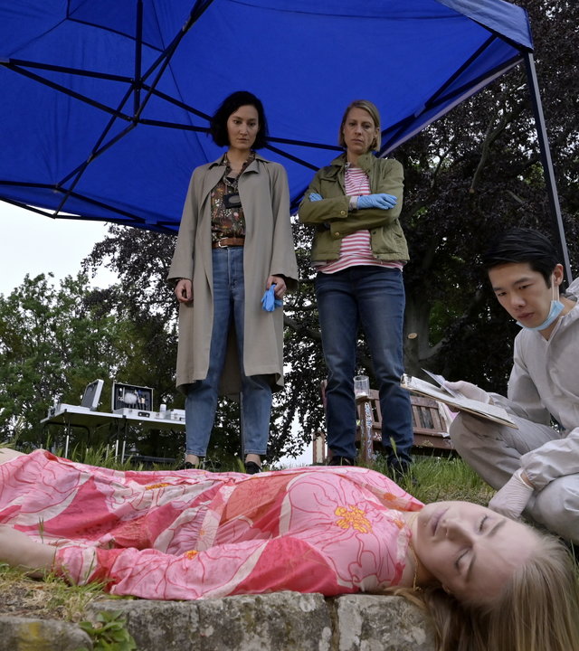 "SOKO Potsdam - Das siebte Haus": Auf einer Wiese auf dem Pfingstberg liegt eine weibliche Leiche, vor der Spurensicherer Thomas Brandner (Yung Ngo) hockt und die Leiche inspiziert. Pauline Hobrecht (Agnes Decker) und Tamara Meurer (Anja Pahl) stehen vor der Leiche unter einem blauen Sonnenschirm und blicken hinunter auf die Leiche.