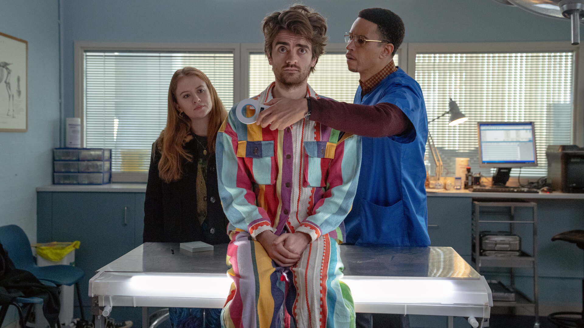 "Extraordinary - Projekt Haustier": In einem Arztzimmer sitzt Jizzlord (Luke Rollason) auf einem Behandlungstisch und blickt skeptisch geradeaus. Hinter ihm steht Dr. Wenderburn (John Macmillan), der einen Metall-Detektor vor seine Brust hält. Jen (Máiréad Tyers) steht ebenfalls hinter Jizzlord und betrachtet ihn nachdenklich.