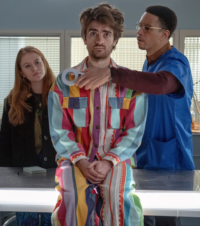 "Extraordinary - Projekt Haustier": In einem Arztzimmer sitzt Jizzlord (Luke Rollason) auf einem Behandlungstisch und blickt skeptisch geradeaus. Hinter ihm steht Dr. Wenderburn (John Macmillan), der einen Metall-Detektor vor seine Brust hält. Jen (Máiréad Tyers) steht ebenfalls hinter Jizzlord und betrachtet ihn nachdenklich.