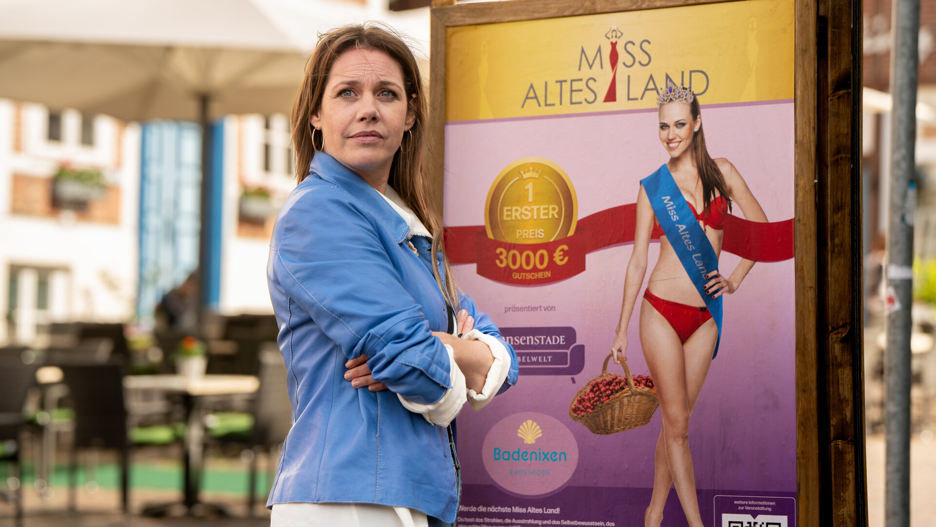 "Neuer Wind im Alten Land - Miss Altes Land": In der Stadt: Beke (Felicitas Woll) steht vor dem Werbeplakat für die Wahl zur "Miss Altes Land". Sie hat die Arme verschränkt und blickt zur Seite.