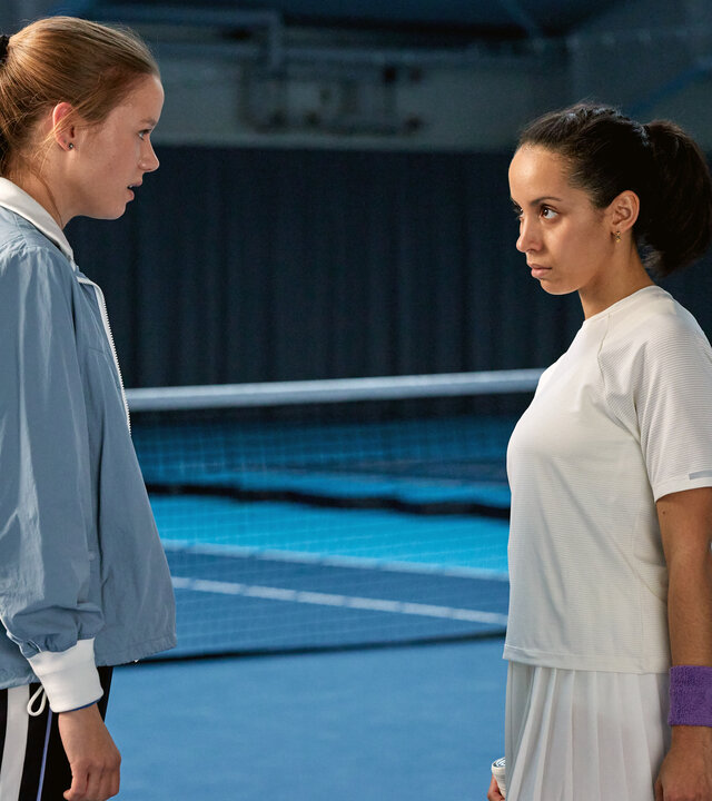"SOKO Hamburg - Der letzte Aufschlag": Vika Antonescu (Lilly Barshy) und Sarah Tulasi (Kristin Alia Hunold) stehen sich in einer Tennishalle auf dem Spielfeld vor dem Netz gegenüber. Sie blicken sich ernst in die Augen, die Körperhaltung der beiden ist angespannt.