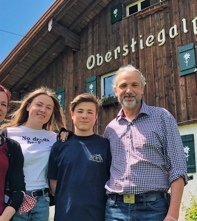 "Terra Xpress: Neustart auf der Alm und Gefahr unterm Gipfelkreuz": Eine Familie, Vater, Mutter, Sohn und Tochter stehen vor einem alten Bauernhaus auf einer Wiese. Darauf steht "Oberstiegalpe".