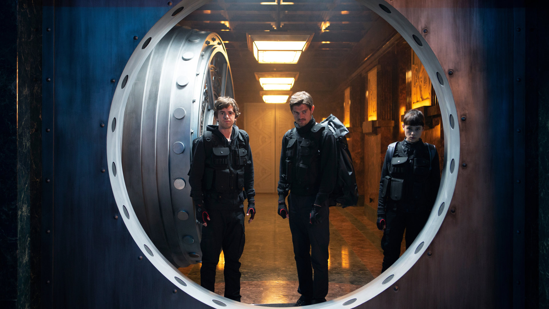 "Countdown in Madrid": Halbtotale. Thom (Freddie Highmore), James (Sam Riley) und Lorraine (Àstrid Bergès-Frisbey) stehen mit angespannten Blicken in offener Tür von Tresorraum. Die Kamera blickt aus dem Tresorraum heraus auf das Trio.