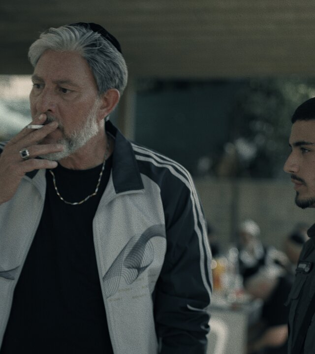 "Borders - Insider": Chicko (Haim Znati) und Kobi (Ido Elieli) stehen nebeneinander im Eingangsbereich eines Hauses. Chicko zieht an einer Zigarette, Kobi blickt in seine Richtung.