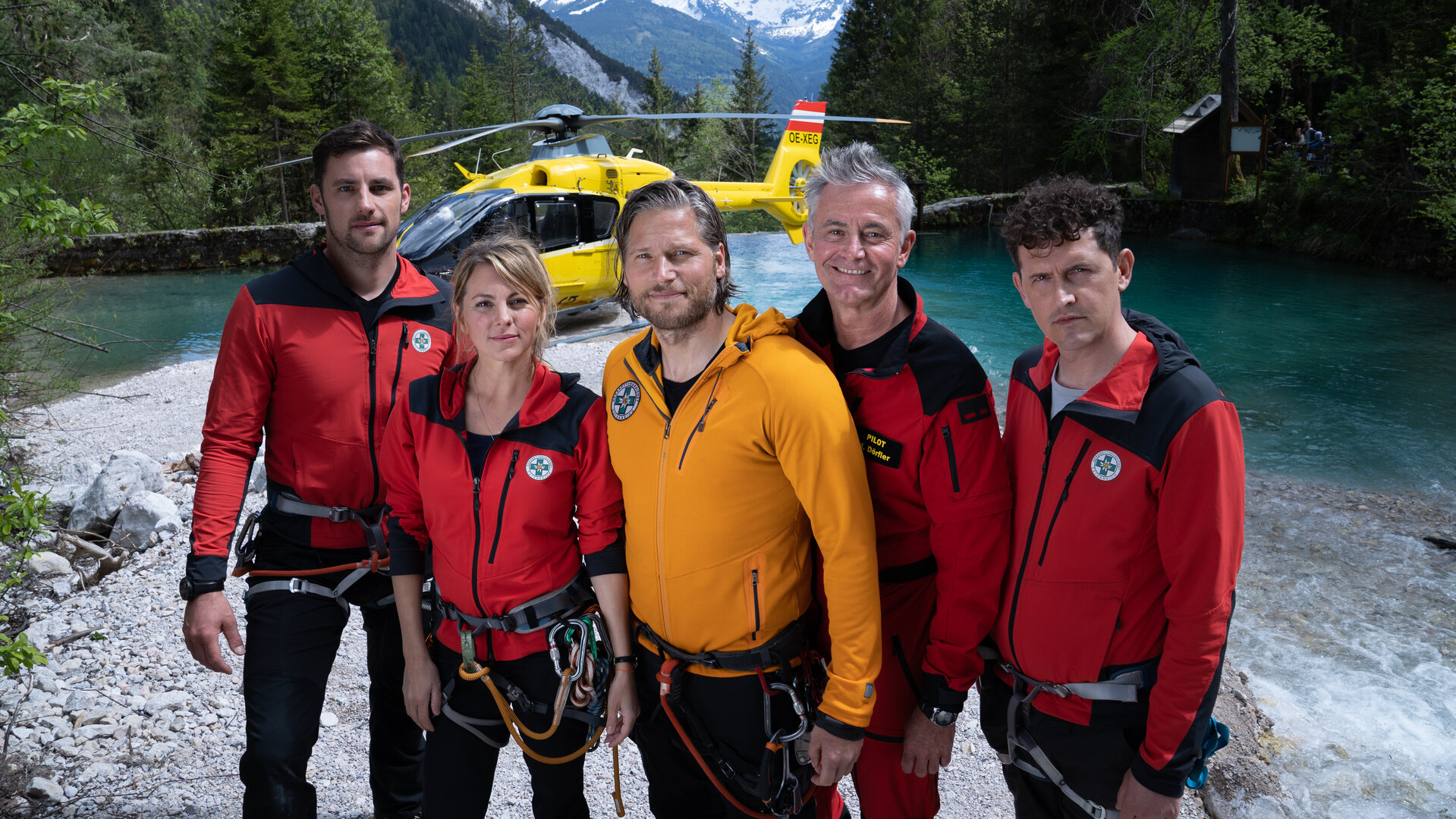 "Die Bergretter: Verbrannte Erde": Gruppenbild des Bergretter-Teams: Markus (Sebastian Ströbel), Katharina (Luise Bähr), Simon (Ferdinand Seebacher), Rudi (Michael Pascher) und Michi (Robert Lohr). Im Hintergrund steht der Rettungshubschrauber vor einer imposanten Bergkulisse.