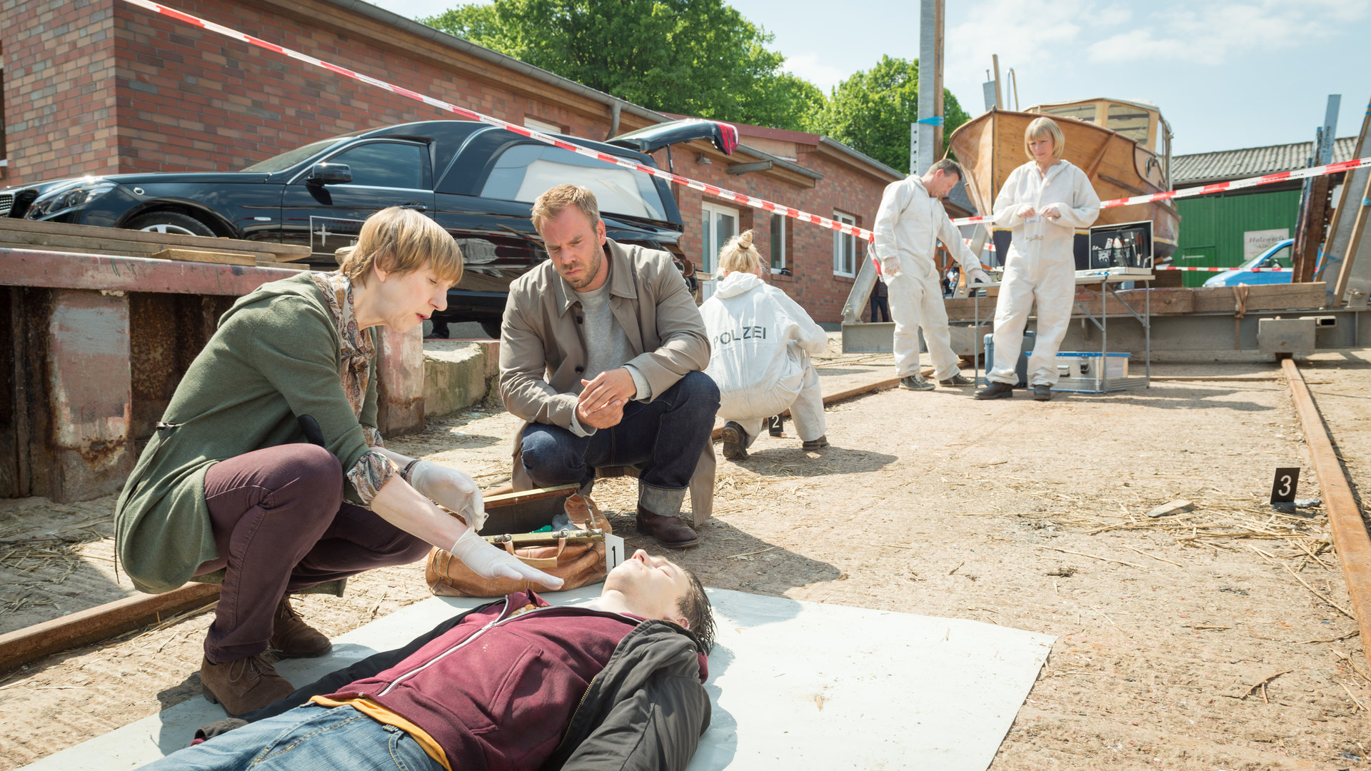 "SOKO Wismar – Tödliche Lichter": Am abgesperrten Tatort hocken Helene Sturbeck (Katharina Blaschke) und Kommissar Lars Pöhlmann (Dominic Boeer) neben der Leiche von Jonas Friese (Komparse). Im Hintergrund sind Roswitha Prinzler (Silke Matthias) und ihre Kollegen (Komparsen) von der KTU bei der Arbeit. Außerdem steht ein schwarzer Leichenwagen mit geöffnetem Kofferraum bereit.