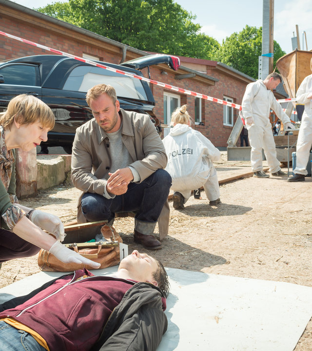 "SOKO Wismar – Tödliche Lichter": Am abgesperrten Tatort hocken Helene Sturbeck (Katharina Blaschke) und Kommissar Lars Pöhlmann (Dominic Boeer) neben der Leiche von Jonas Friese (Komparse). Im Hintergrund sind Roswitha Prinzler (Silke Matthias) und ihre Kollegen (Komparsen) von der KTU bei der Arbeit. Außerdem steht ein schwarzer Leichenwagen mit geöffnetem Kofferraum bereit.