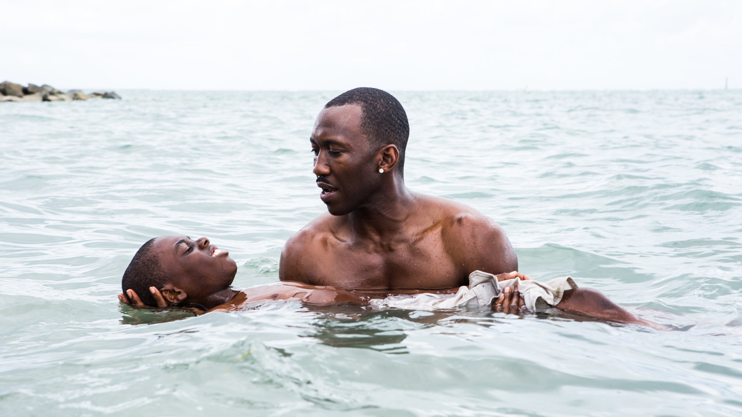 "Moonlight": Ein muskulöser Mann afroamerikanischer Herkunft steht mit nacktem Oberkörper im Wasser und unterstützt auf seinen ausgebreiteten Armen vorsichtig einen Jungen, der mit dem Rücken auf der Wasseroberfläche liegt.