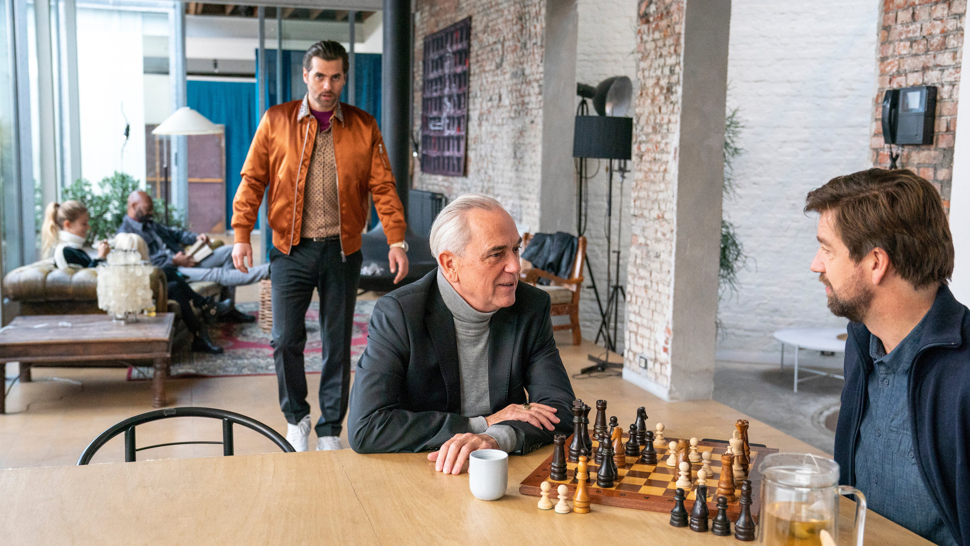 "The Bank Hacker": Alidor Van Praet (Gene Bervoets) spielt Schach mit Bart (Koen De Graeve) an einem Tisch, während Prince (Manuel Broekman) durch den Raum zu ihnen läuft. Im Hintergrund sitzen Ada (Ella-June Henrard) und Souleymane (Claude Musungayi) auf dem Sofa.