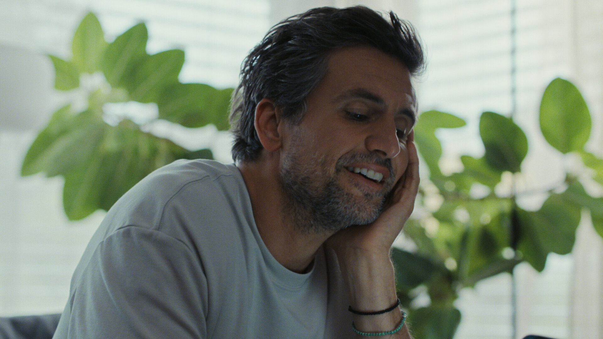 "The other gAIrl – Frag mich, wie’s mir geht": Frank (Tom Beck) sitzt auf dem Sofa und stützt den Kopf in die Hand. Er lächelt. Im Hintergrund stehen Pflanzen auf der Fensterbank.