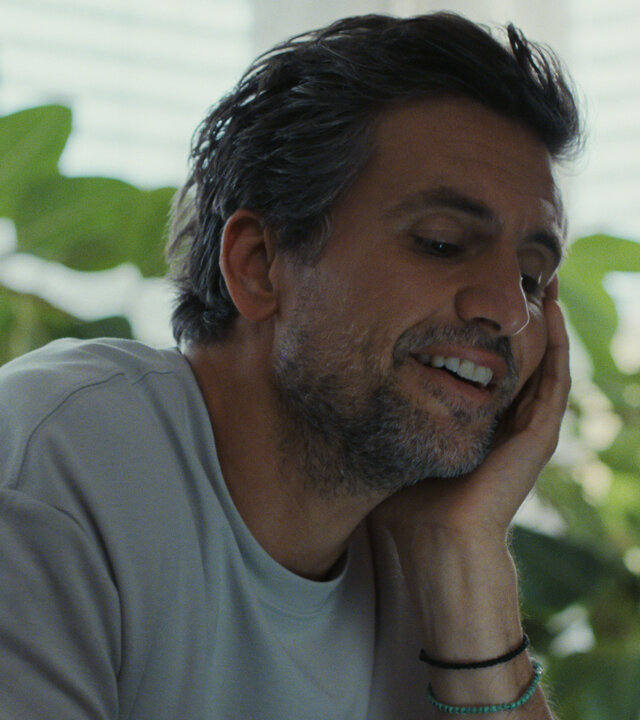 "The other gAIrl – Frag mich, wie’s mir geht": Frank (Tom Beck) sitzt auf dem Sofa und stützt den Kopf in die Hand. Er lächelt. Im Hintergrund stehen Pflanzen auf der Fensterbank.