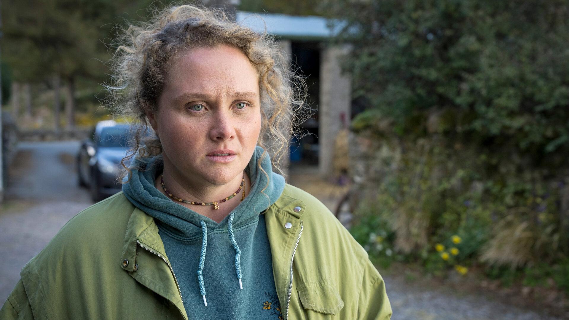 "The Tourist - Irisches Blut - Der Gang der Dinge": Helen (Danielle Macdonald) steht in der Einfahrt zu einem Anwesen und schaut zur Seite, hinter ihr steht ein Taxi. Sie trägt einen azurblauen Hoodie und darüber eine hellgrüne Jacke.