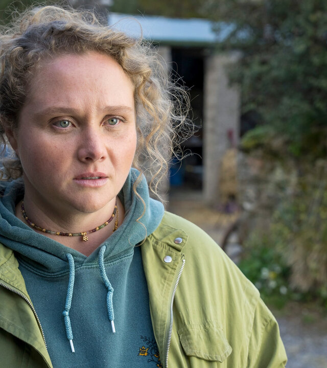"The Tourist - Irisches Blut - Der Gang der Dinge": Helen (Danielle Macdonald) steht in der Einfahrt zu einem Anwesen und schaut zur Seite, hinter ihr steht ein Taxi. Sie trägt einen azurblauen Hoodie und darüber eine hellgrüne Jacke.