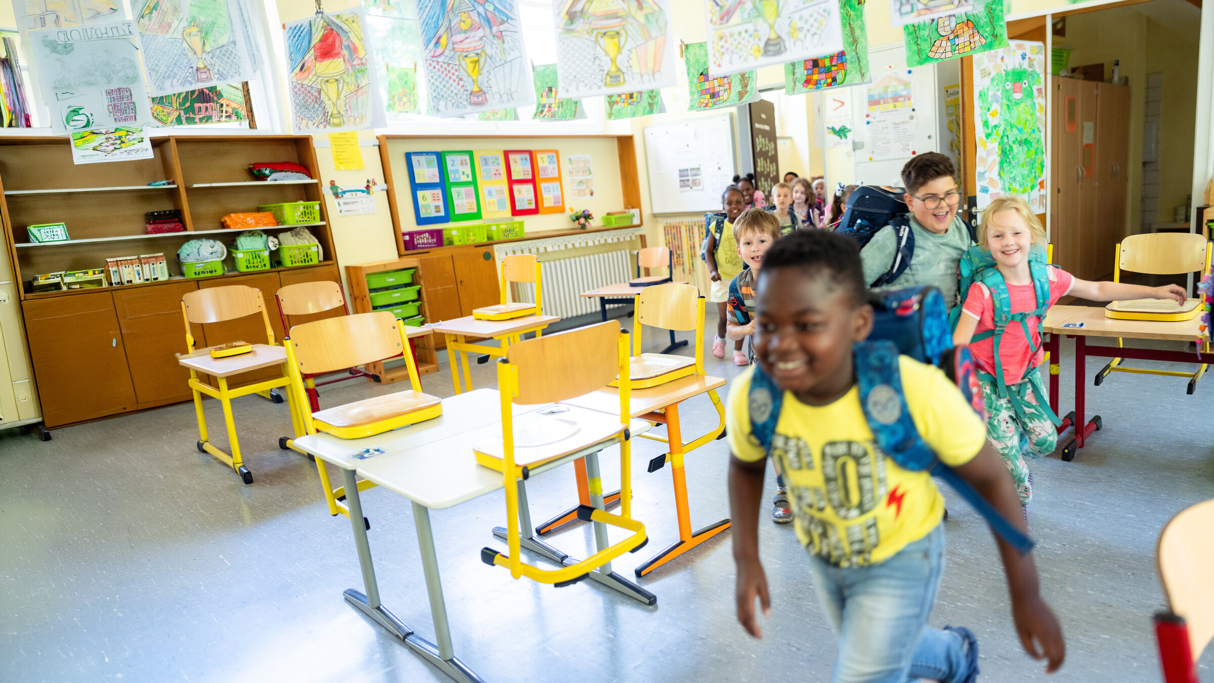 "37Â°: Unsere Klasse: Chaos und Chancen in der 1d": SchÃ¼ler rennen mit RucksÃ¤cken auf dem RÃ¼cken lÃ¤chelnd in einer Reihe zwischen den Tischen durch in ein Klassenzimmer hinein.