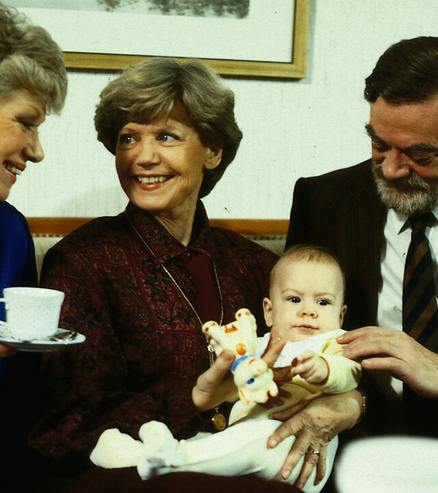 "Die Wicherts von nebenan: Sport ist gesund": Graf (Friedrich Schütter) und Gräfin von Strelenau (Gisela Peltzer) sitzen neben Hannelore (Maria Sebaldt) auf dem Sofa. Die Gräfin hält Baby Katharina auf dem Schoß und lächelt Hannelore an.