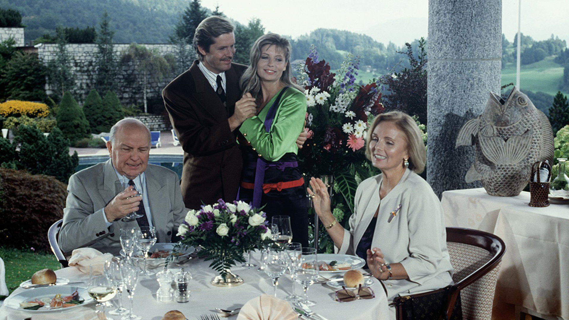 "Das Erbe der Guldenburgs - Die wahre Liebe": Margot (Ruth Maria Kubitschek) und Horst Strobel (Wolfgang Wahl) sitzen am Tisch und speisen. Jan (Sigmar Solbach) und Barbara (Sabine Postel) stehen eng umschlungen neben dem Tisch.