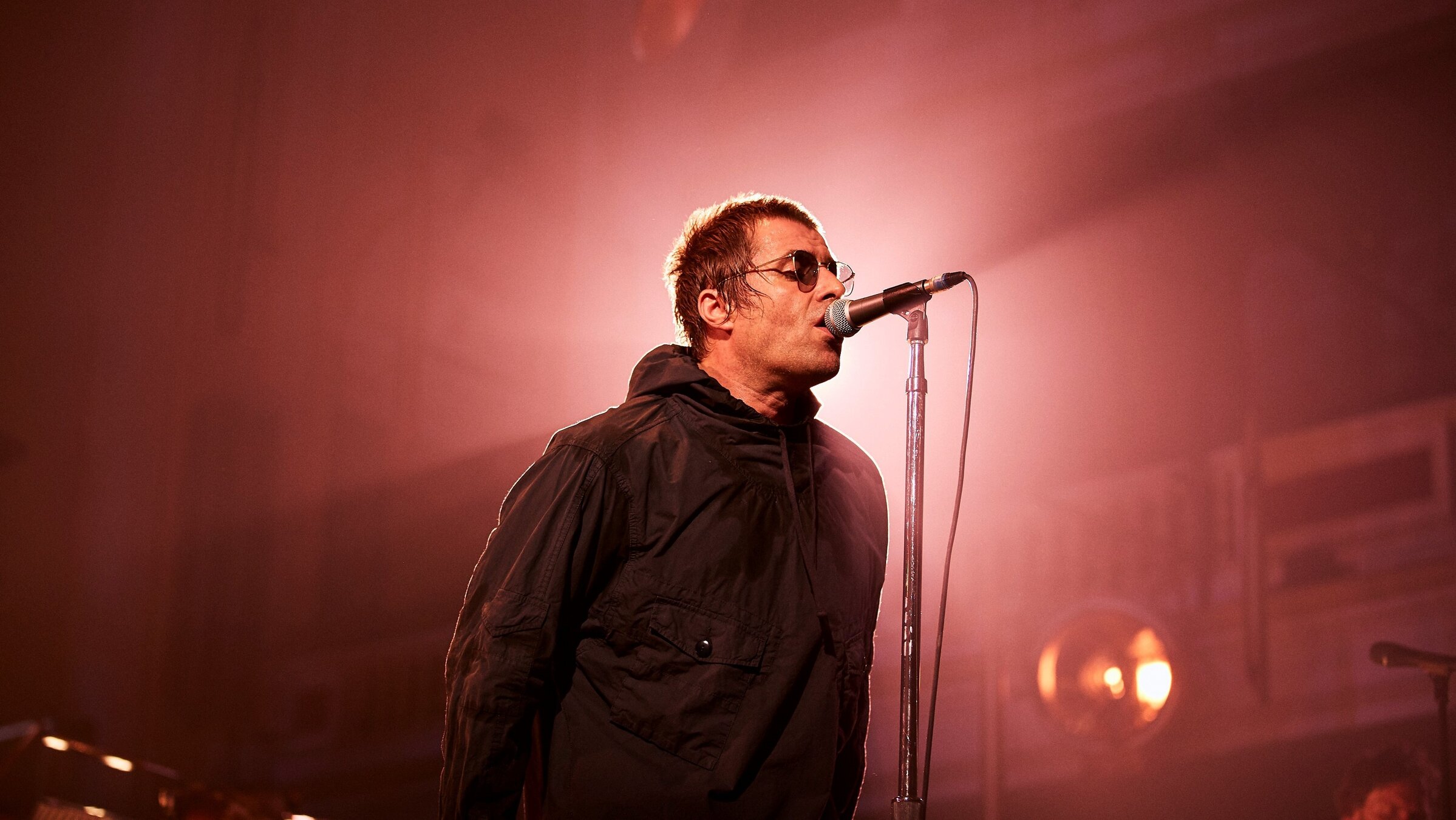 "Liam Gallagher: Live at Knebworth 2022": Der Sänger Liam Gallagher steht seitlich vor einem Mikrofon und singt. Er trägt eine schwarze Jacke mit Kapuze und eine Sonnenbrille. Der Raum um ihn herum ist in ein diffuses Licht getaucht, Details kaum erkennbar.