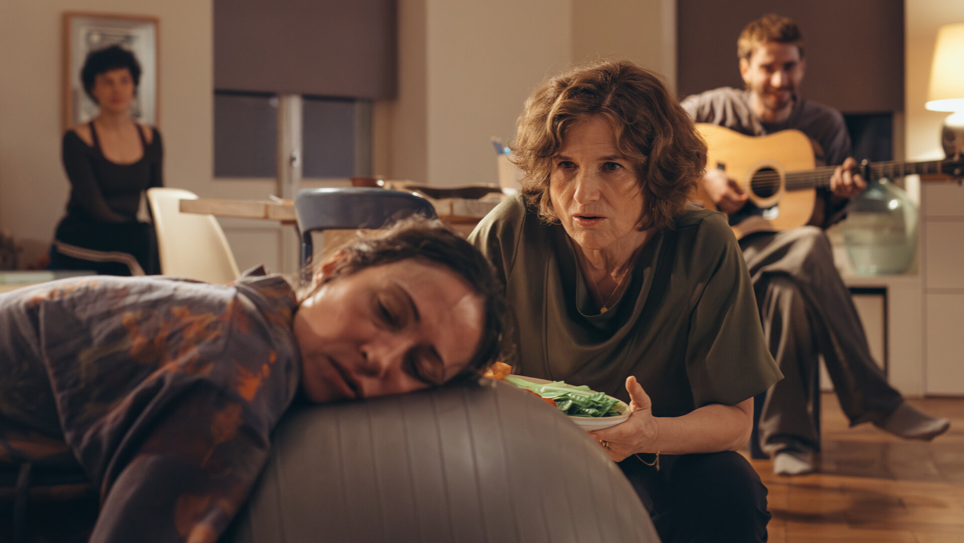 "PUSH - Trust the process": Raquel (Aenne Schwarz) liegt erschöpft mit ihrem Kopf auf einem Gymnastikball. Marcela (Victoria Trauttmansdorff) kniet neben ihr mit einem Gemüseteller und spricht mit Raquel. Leandro (James Newton) sitzt dahinter und spielt Gitarre. Greta (Lydia Amasko) sitzt im Hintergrund.