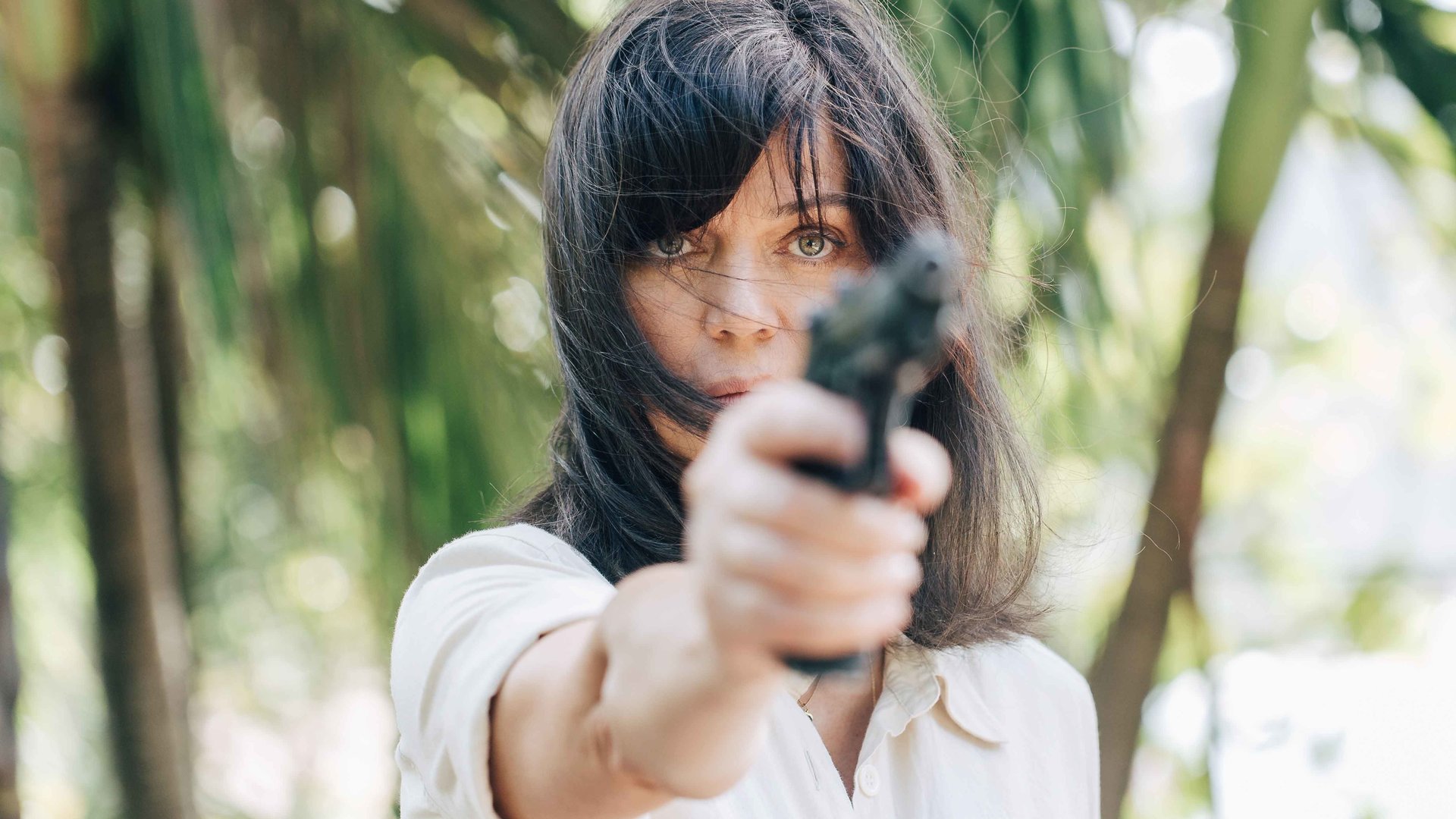 "They Were Ten": Eve (Marianne Denicourt) hält mit ausgestrecktem rechten Arm eine Pistole nach oben. Im Hintergrund sind unscharf Palmen zu sehen.