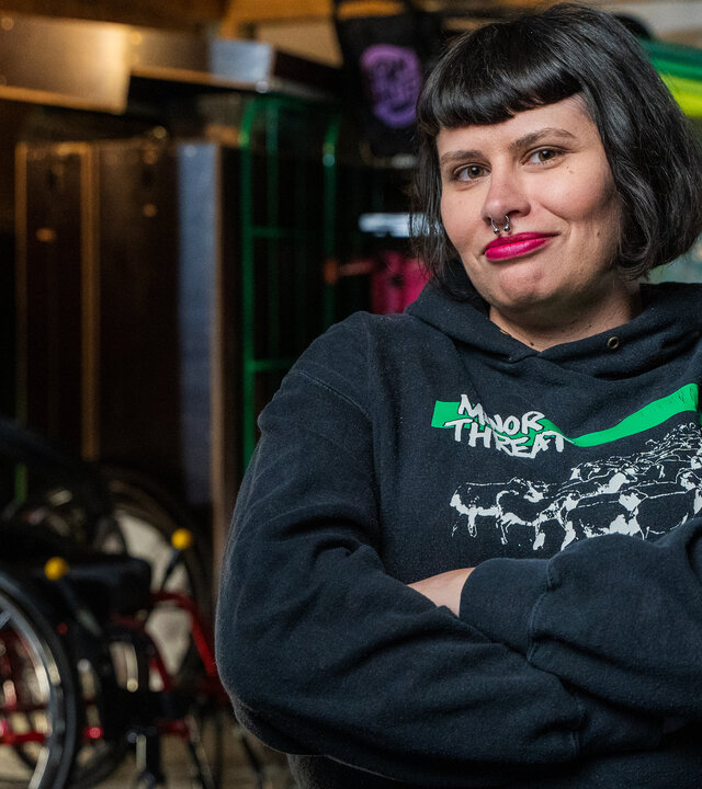 "einfach Mensch - Lisa Lebuser: Im Rollstuhl Grenzen sprengen": Die Protagonistin Lisa Lebuser sitzt in einem Raum und schaut lächelnd in die Kamera. Im Hintergrund stehen mehrere Rollstühle.