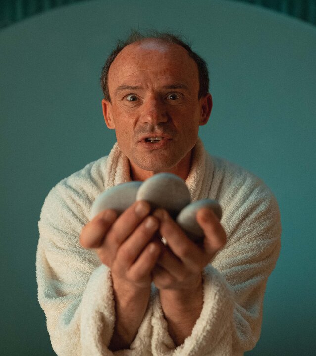 "Schlechte Menschen - Der Nörgler": Gustav (Gerhard Liebmann) hält drei flache Steine anbietend vor sich und sieht intensiv in die Kamera.