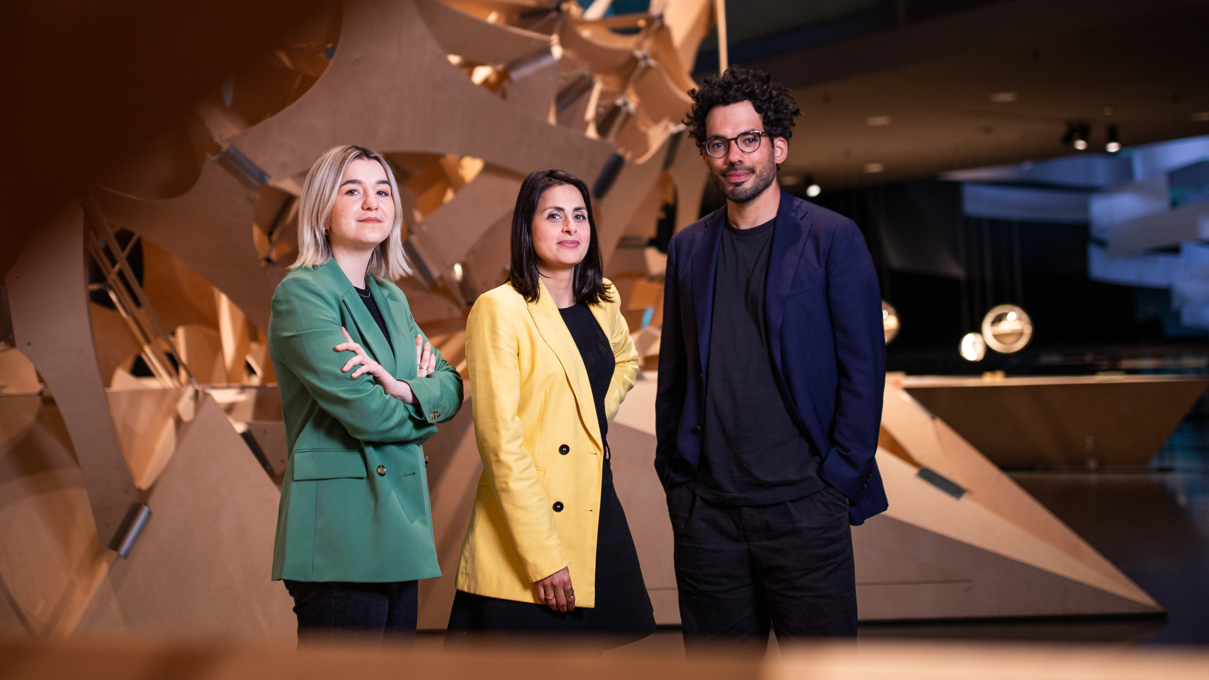 "Futur Wir – Das neue deutsche Selbstverständnis (3/3) - Wie radikal wir sein wollen": Die drei Moderatoren stehen im Berliner Futurium und blicken in die Kamera. Eingerahmt sind sie von einer großen Holzskulptur.