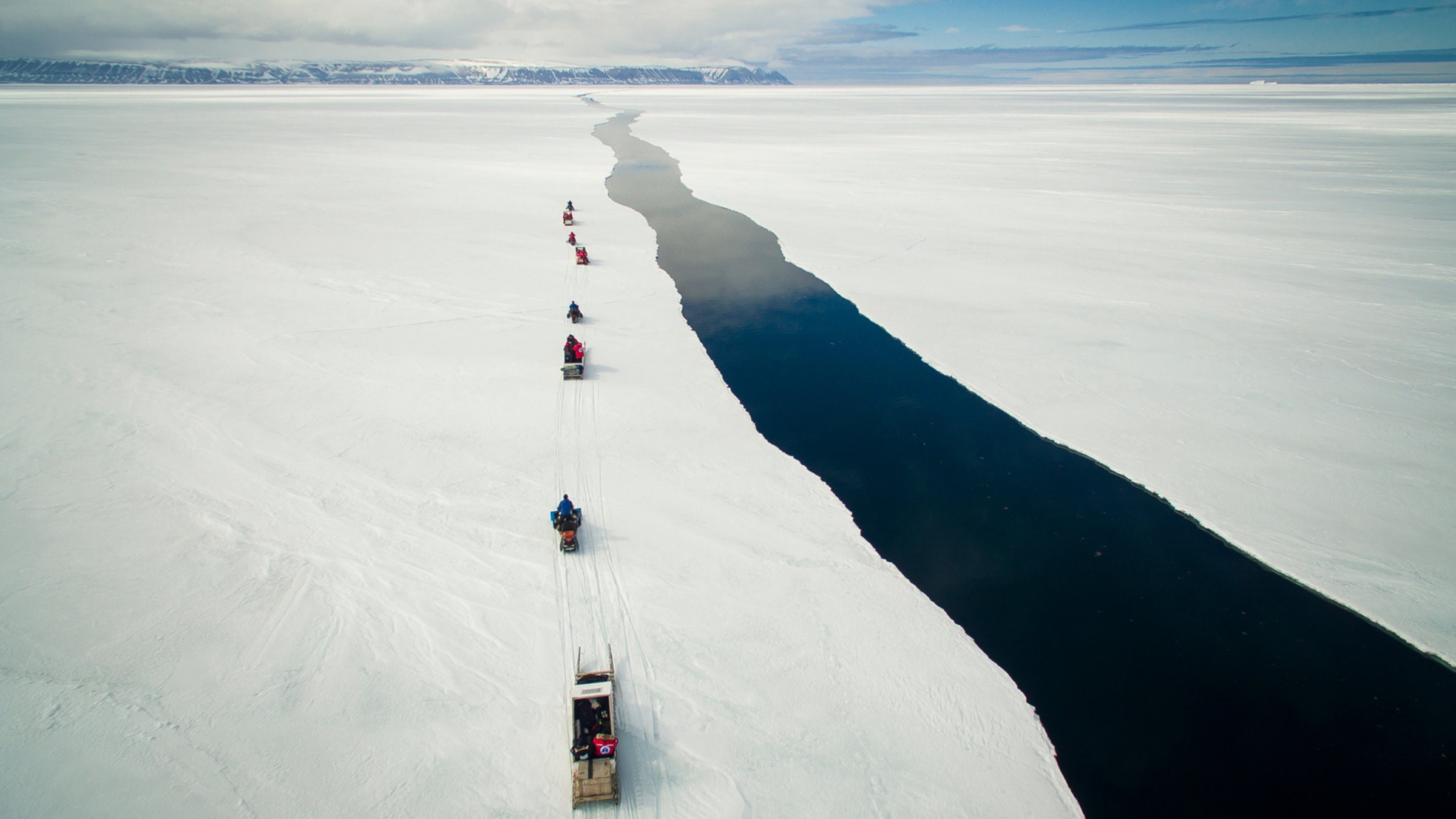"Schutzprojekt Erde": Weite offene Fläche eines zugefrorenen Gewässers. Quer durchs Bild eine Fahrrinne. Links davon eine Karawane mit Lastenschlitten. Am Horizont eine Bergkette.