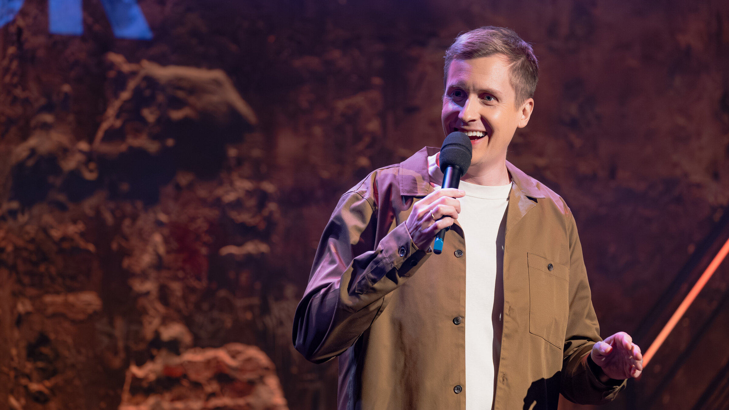 "Till Reiners` Happy Hour": Der Moderator und Stand-Up-Comedian Till Reiners steht mit einem Mikrofon in der Hand auf der Bühne und spricht zum Publikum.