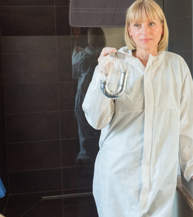 "SOKO Wismar – Tödliche Nähe": Roswitha Prinzler (Silke Matthias) steht in Arbeitskleidung in einem Badezimmer und hält einen in Schutzhülle verpackten abmontierten Siphon hoch. Katrin Börensen (Claudia Schmutzler) und Lars Pöhlmann (Dominic Boeer) blicken in ihre Richtung.