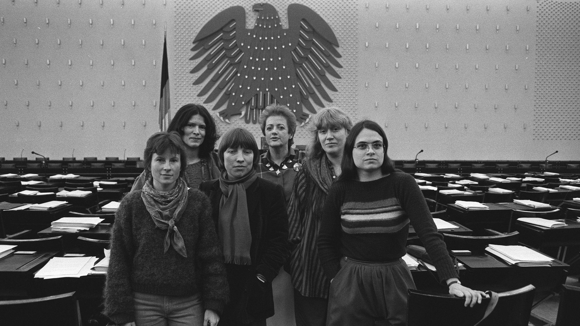 "Die Unbeugsamen": SW-Fotos von 1984: eine Gruppe von sechs Frauen steht zusammen, im Hintergrund die leeren Abgeordnetentische und Regierungsband des deutschen Bundestages in Bonn. Es sind die Sprecherinnen der Fraktion der Partei Die Grünen.