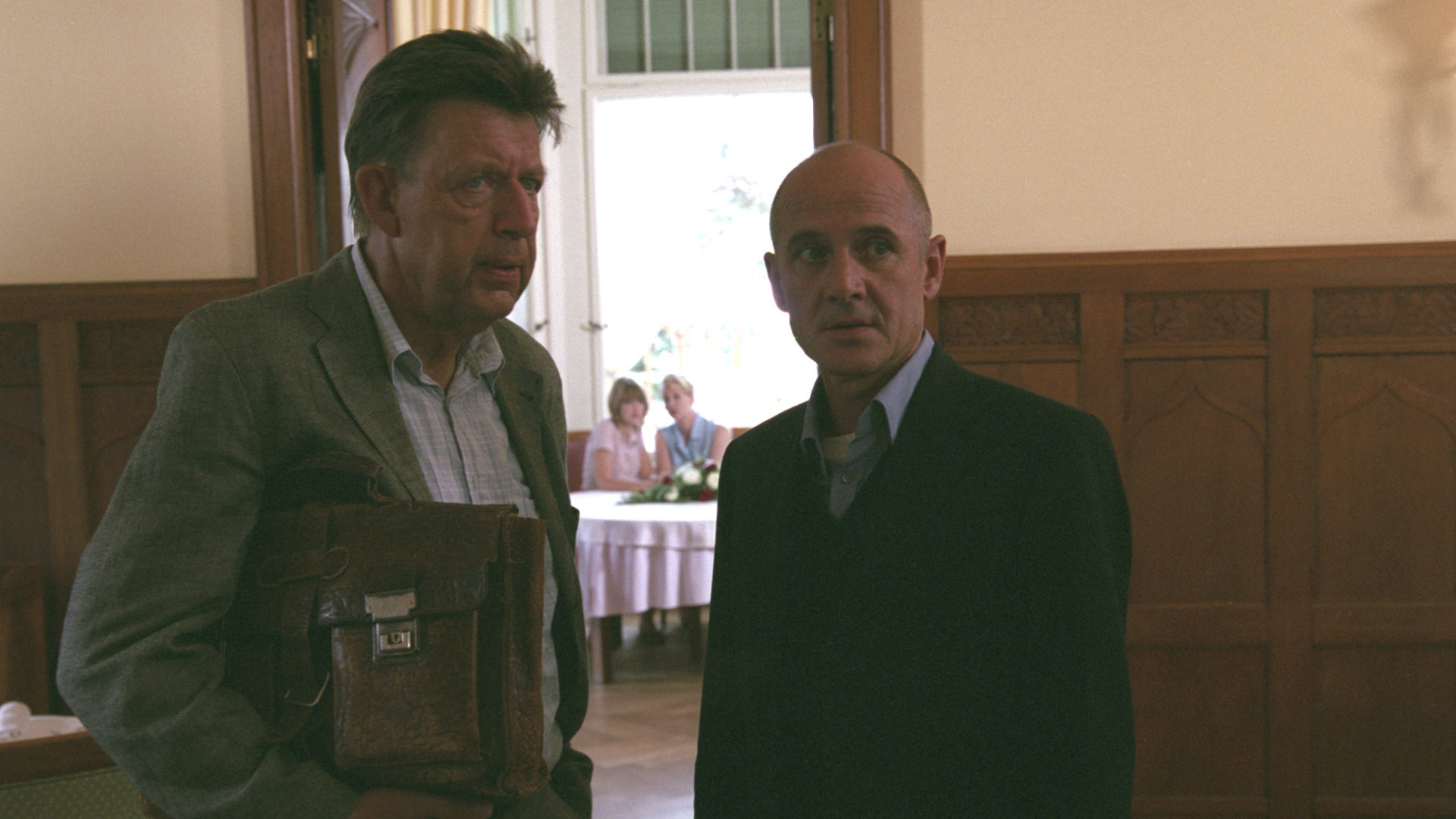 "Der letzte Zeuge - Unter Schwestern": Joe Hoffer (Jörg Gudzuhn) und Dr. Kolmaar (Ulrich Mühe)  stehen nebeneinander in einem großen Raum und beobachten etwas.