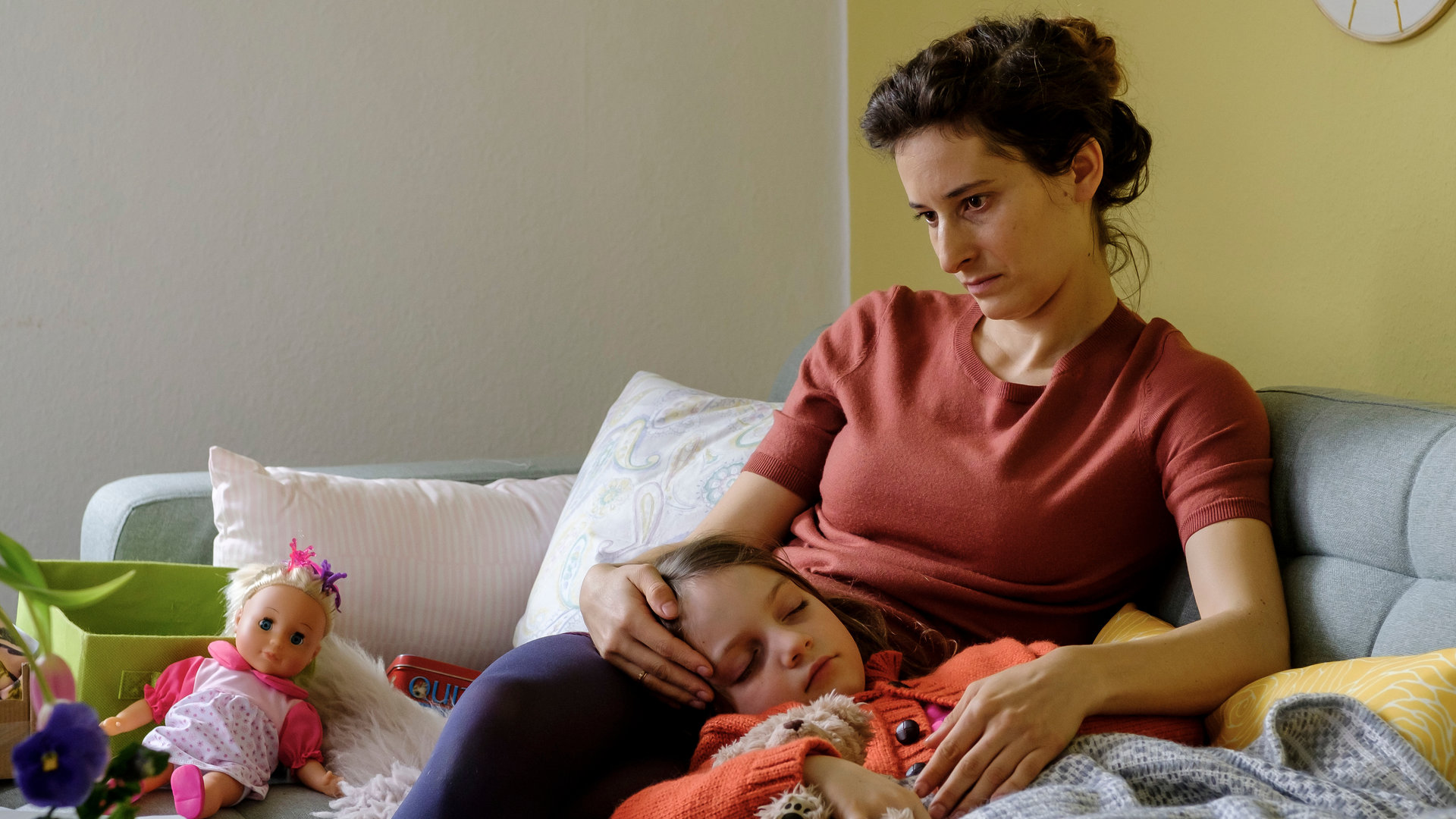 "SOKO Potsdam - Zu deinem Schutz": Carola Blum (Stella Hilb) sitzt auf einem grauen Sofa, auf der ihre kranke und schlafende Tochter Emilia (Rosa Wirtz) liegt. Ihr Kopf liegt auf den Oberschenkeln der Mama, die hoffnungslos zu Boden schaut.