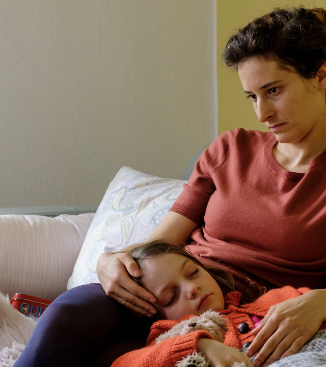 "SOKO Potsdam - Zu deinem Schutz": Carola Blum (Stella Hilb) sitzt auf einem grauen Sofa, auf der ihre kranke und schlafende Tochter Emilia (Rosa Wirtz) liegt. Ihr Kopf liegt auf den Oberschenkeln der Mama, die hoffnungslos zu Boden schaut.